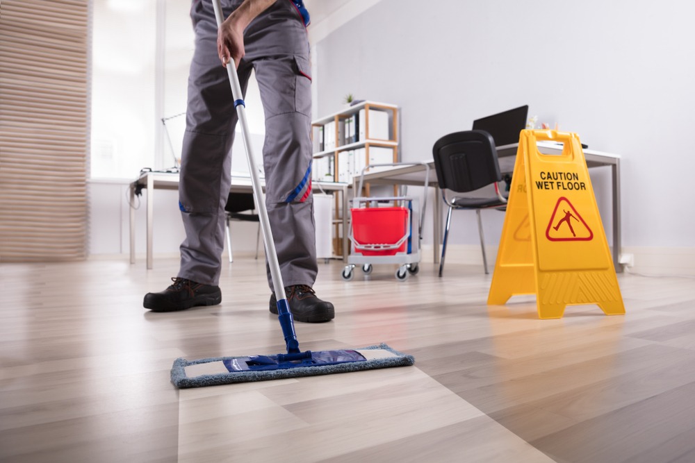 janitor in uniform cleaning floor in room with wood floor and wet floor sign